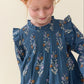 A young girl with red hair looking down, wearing a blue long-sleeve smocked dress with a white and purple floral pattern, against a plain cream background.