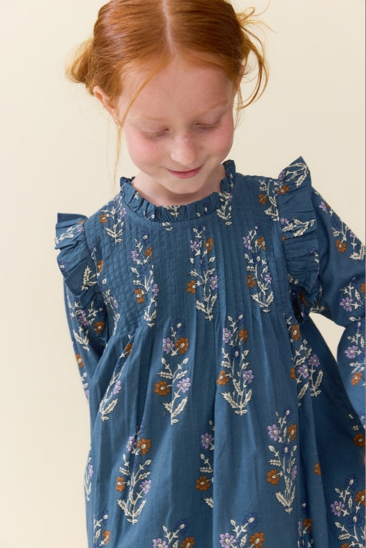 A young girl with red hair looking down, wearing a blue long-sleeve smocked dress with a white and purple floral pattern, against a plain cream background.