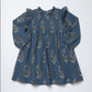 A long-sleeve blue-gray cotton dress with a delicate floral and botanical print in muted brown, purple, and beige tones, featuring ruffled shoulders and a gathered neckline, laid flat against a white background.