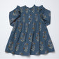 A toddler's long-sleeve dress in slate blue cotton fabric, decorated with a repeating floral pattern in brown, purple, and cream. The dress features a smocked bodice, a button-up front with light blue buttons, and ruffled shoulders, laid flat against a white background.