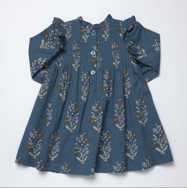 A toddler's long-sleeve dress in slate blue cotton fabric, decorated with a repeating floral pattern in brown, purple, and cream. The dress features a smocked bodice, a button-up front with light blue buttons, and ruffled shoulders, laid flat against a white background.