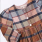A close-up of a children's plaid dress in muted earthy tones, featuring a mix of dusty pink, brown, navy, and beige stripes. The dress has a button-down front, long sleeves with gathered cuffs, and a gathered waist seam.