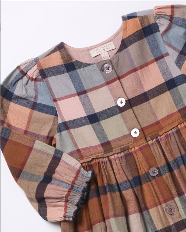 A close-up of a children's plaid dress in muted earthy tones, featuring a mix of dusty pink, brown, navy, and beige stripes. The dress has a button-down front, long sleeves with gathered cuffs, and a gathered waist seam.