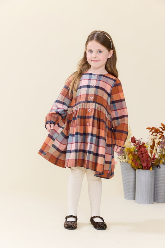 A young girl stands against a warm cream studio background wearing a long-sleeve, tiered plaid dress in rust orange, navy, and beige. She is styled with white tights and dark brown shoes, with decorative flower pots on the side.