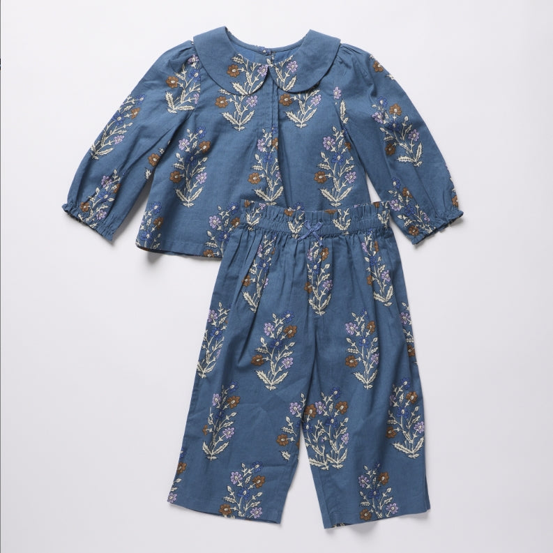 A two-piece children's outfit set consisting of a long-sleeve top with a Peter Pan collar and matching wide-leg pants, both in a dusty blue fabric with a colorful floral print, laid flat on a light gray background.