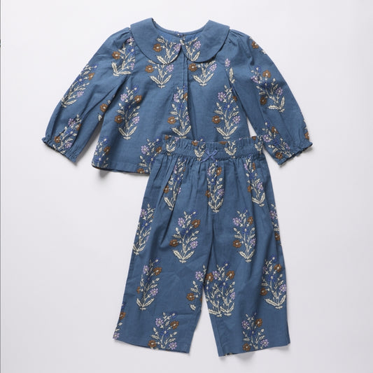 A two-piece children's outfit set consisting of a long-sleeve top with a Peter Pan collar and matching wide-leg pants, both in a dusty blue fabric with a colorful floral print, laid flat on a light gray background.