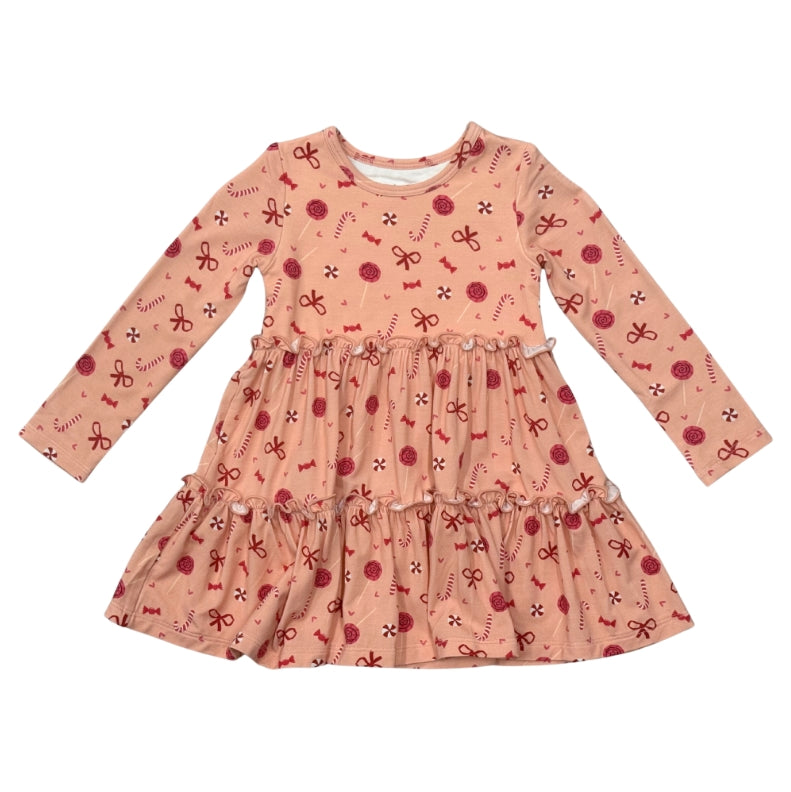 A long-sleeve baby girl's dress in a peachy pink fabric with an all-over red and white candy cane and bow pattern, featuring a smocked waist and a tiered ruffle skirt.
