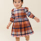A toddler girl wearing a long-sleeve plaid dress in orange, blue, and pink, standing against a plain cream background.