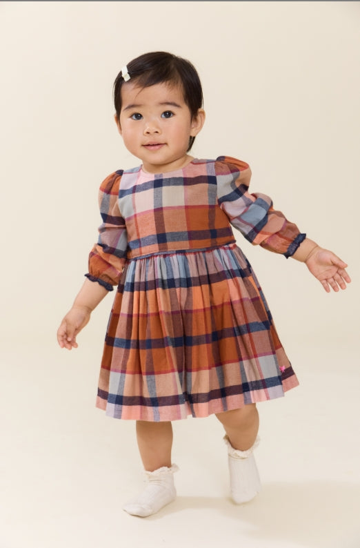A toddler girl wearing a long-sleeve plaid dress in orange, blue, and pink, standing against a plain cream background.