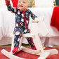 A baby wearing a navy blue Christmas-themed footed pajama set sits on a white wooden rocking horse in a decorated bedroom setting.