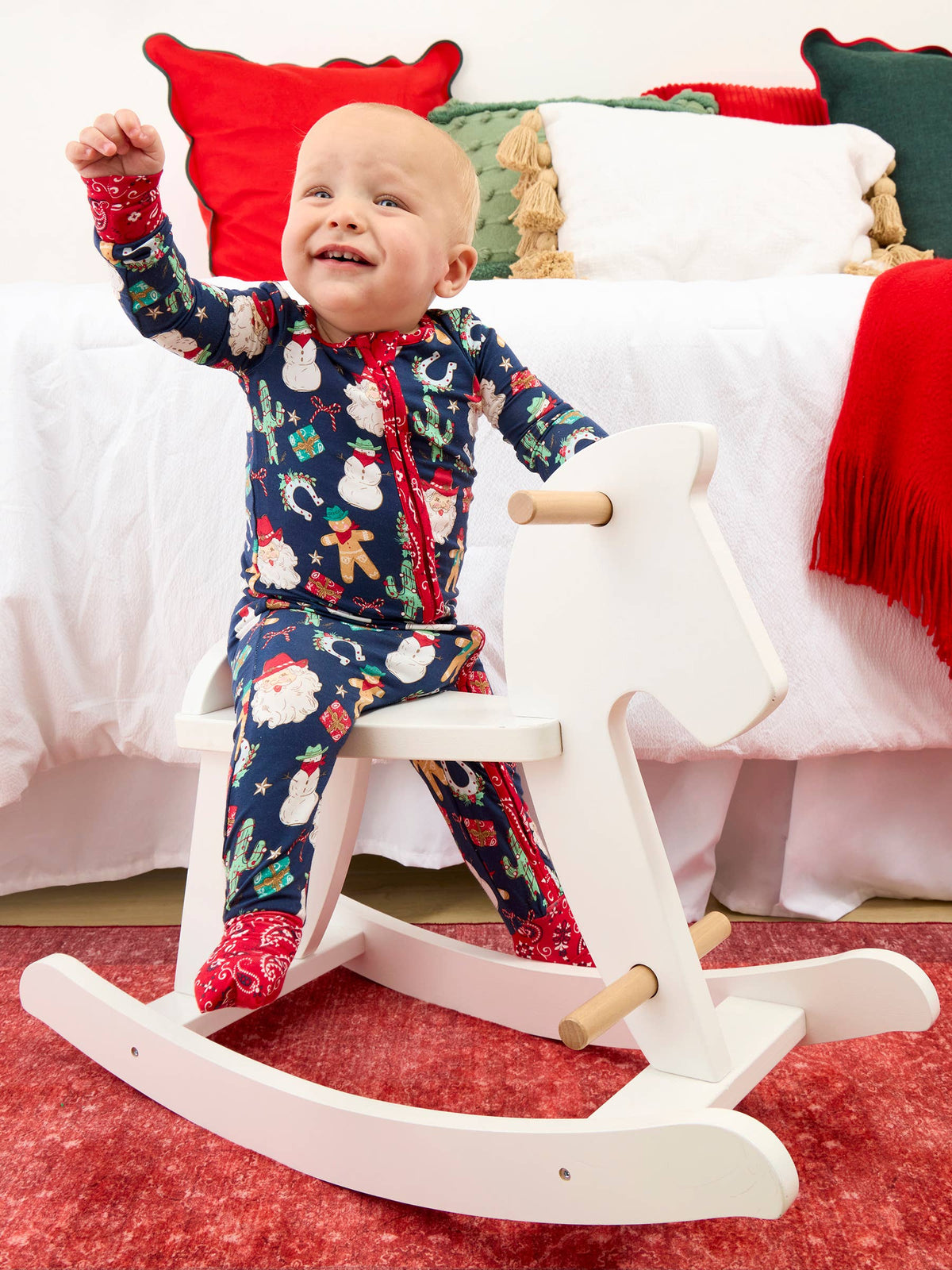 A baby wearing a navy blue Christmas-themed footed pajama set sits on a white wooden rocking horse in a decorated bedroom setting.