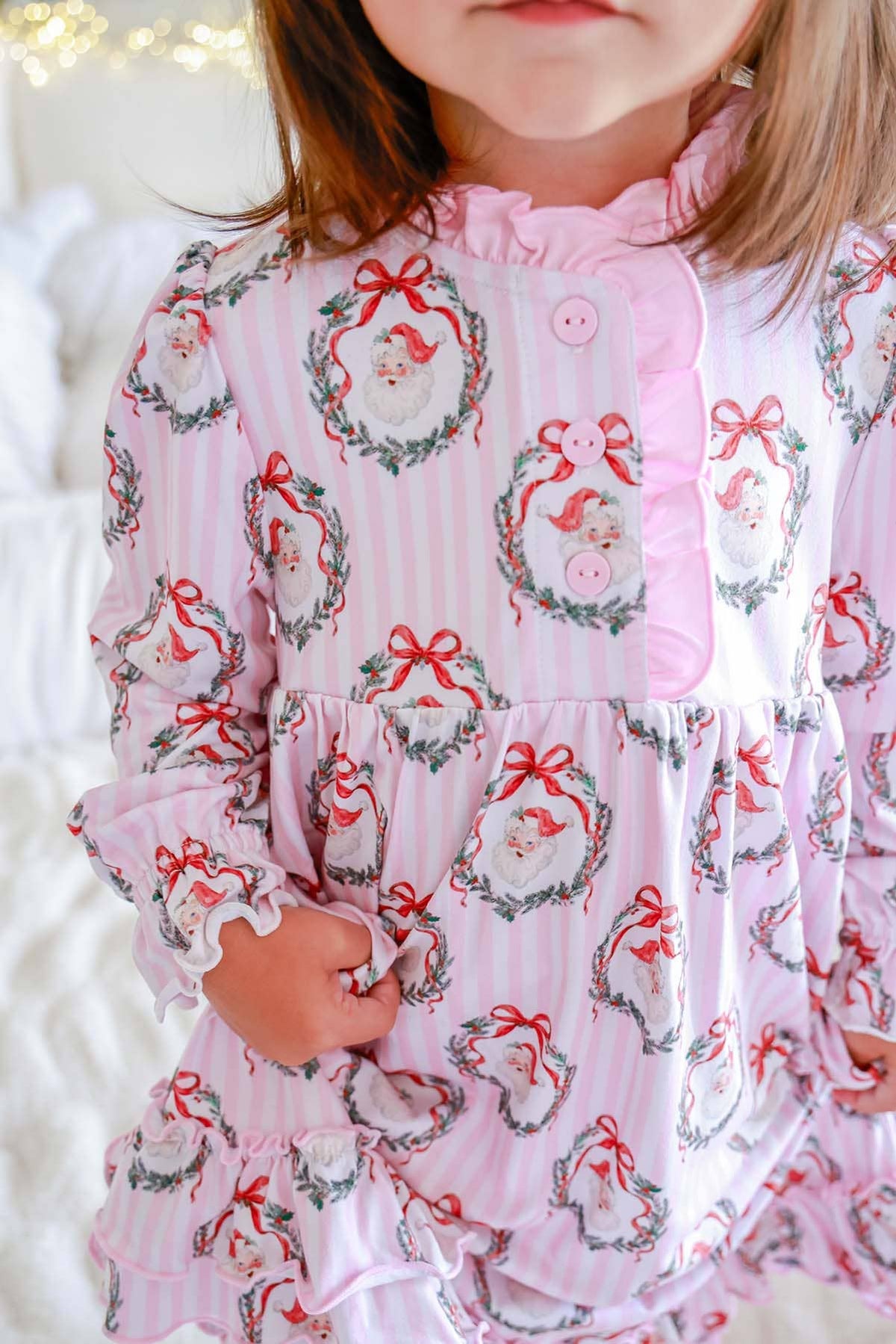 A close-up of a child wearing a pink long-sleeve pajama top with a festive Santa Claus wreath pattern, featuring ruffled trim and a button placket.