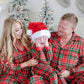 A family of three wearing matching red and green plaid Christmas pajamas sits together on a carpeted floor in front of decorated Christmas trees and stockings. The child in the center wears a Santa hat and covers their face with their hands.