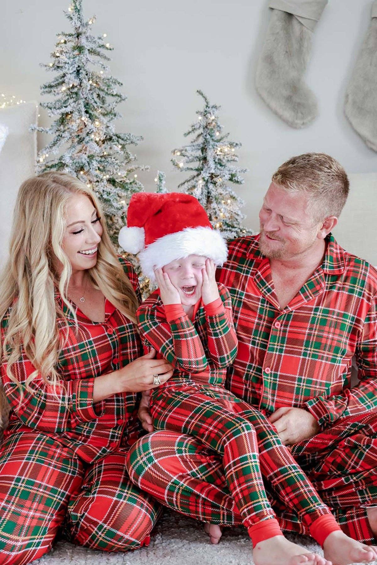 A family of three wearing matching red and green plaid Christmas pajamas sits together on a carpeted floor in front of decorated Christmas trees and stockings. The child in the center wears a Santa hat and covers their face with their hands.