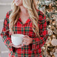 A woman wearing a red and green plaid pajama set holds a white mug in front of a decorated Christmas tree.