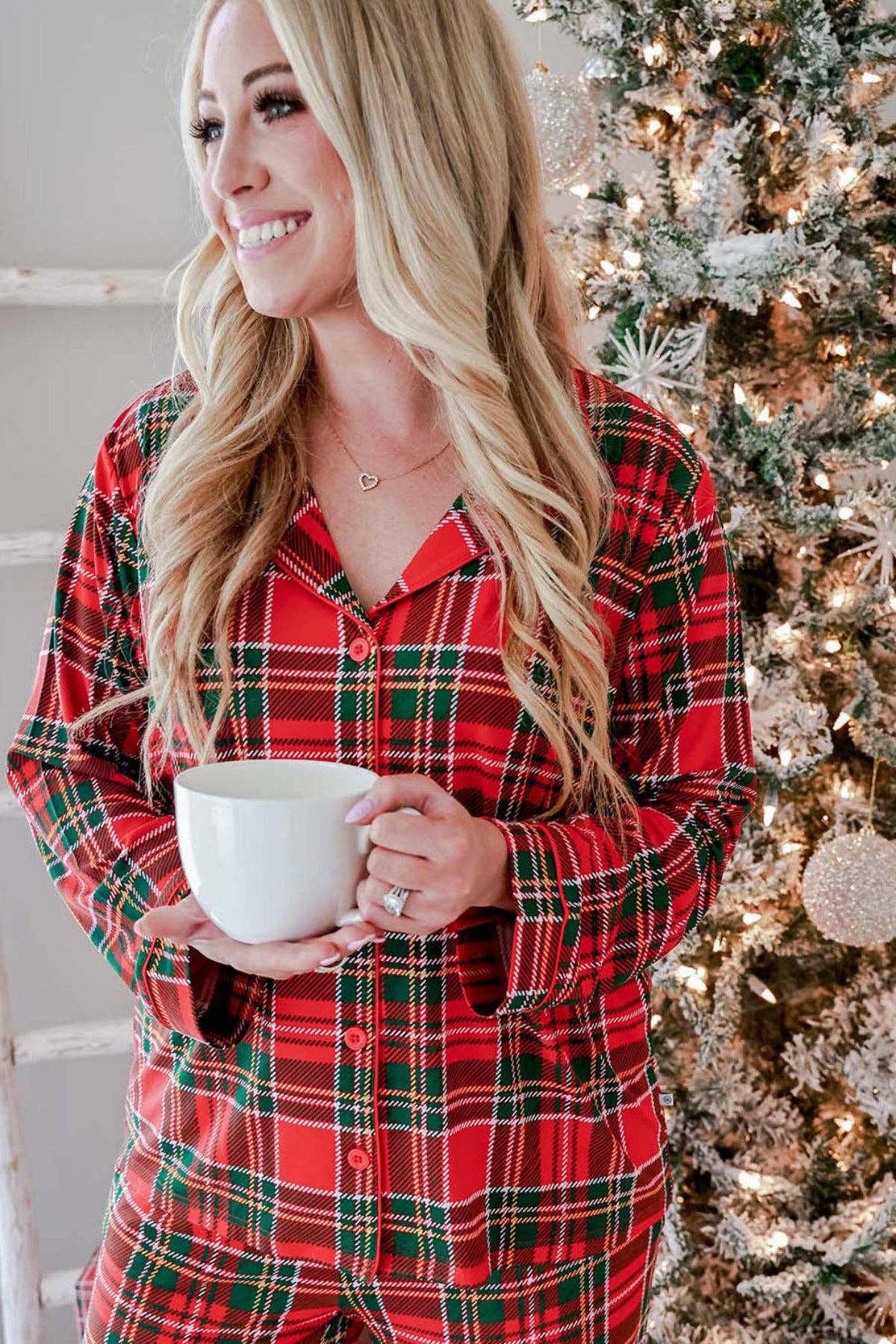 A woman wearing a red and green plaid pajama set holds a white mug in front of a decorated Christmas tree.