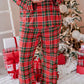 A person wearing a red, green, and black plaid pajama set stands barefoot in a festive holiday setting, holding a white mug near a decorated Christmas tree and wrapped gifts.