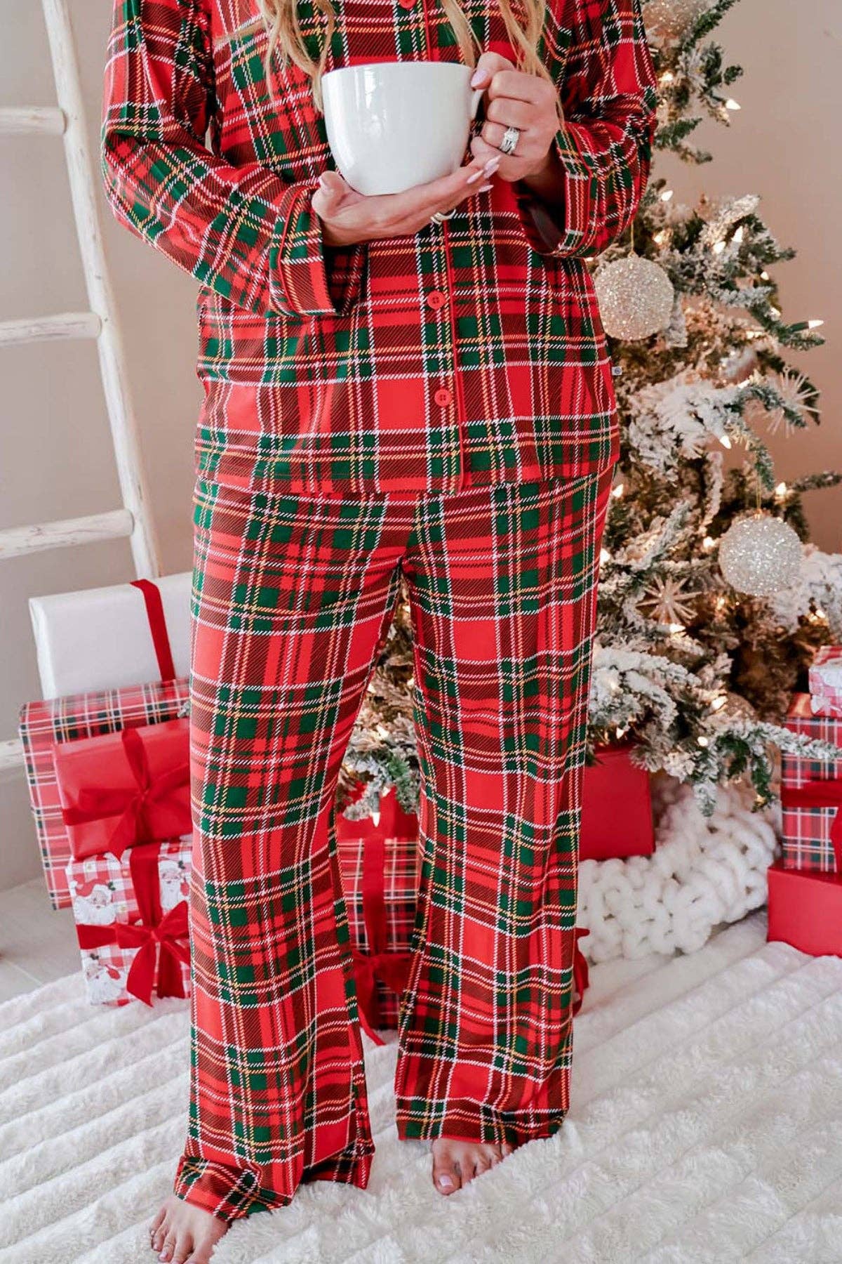 A person wearing a red, green, and black plaid pajama set stands barefoot in a festive holiday setting, holding a white mug near a decorated Christmas tree and wrapped gifts.