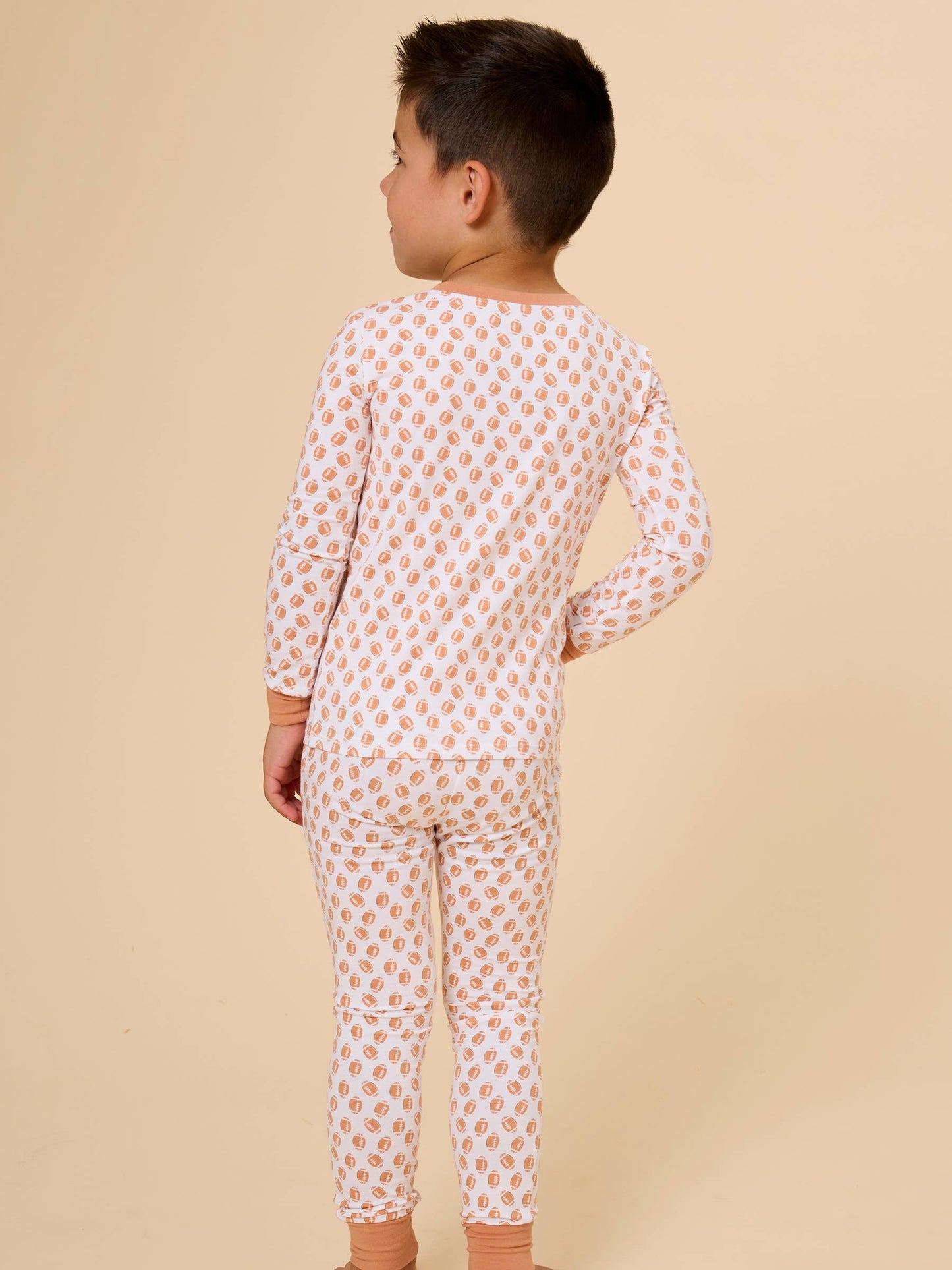 A young boy viewed from the back wearing a long-sleeve pajama set with a small orange and white repeating pattern, set against a solid beige studio background.