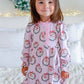 A young girl with a pink bow in her hair stands on a white bed wearing a pink long-sleeve nightgown with a festive holiday wreath and snowman print. The background features a white wall, a green wreath, and warm string lights.