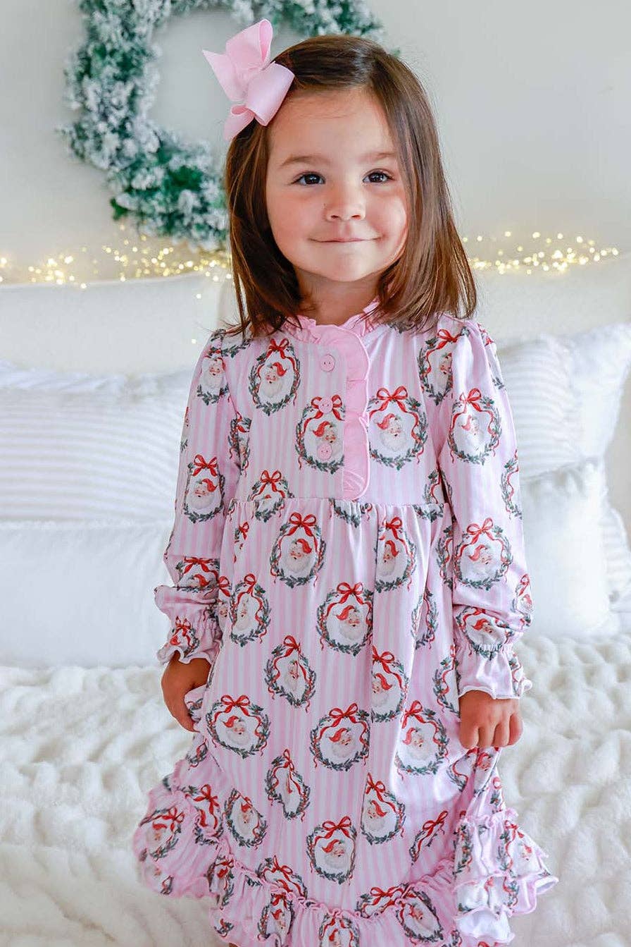 A young girl with a pink bow in her hair stands on a white bed wearing a pink long-sleeve nightgown with a festive holiday wreath and snowman print. The background features a white wall, a green wreath, and warm string lights.