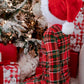 A child wearing red and green plaid pajamas and a Santa hat sits in front of a decorated Christmas tree with wrapped presents nearby.