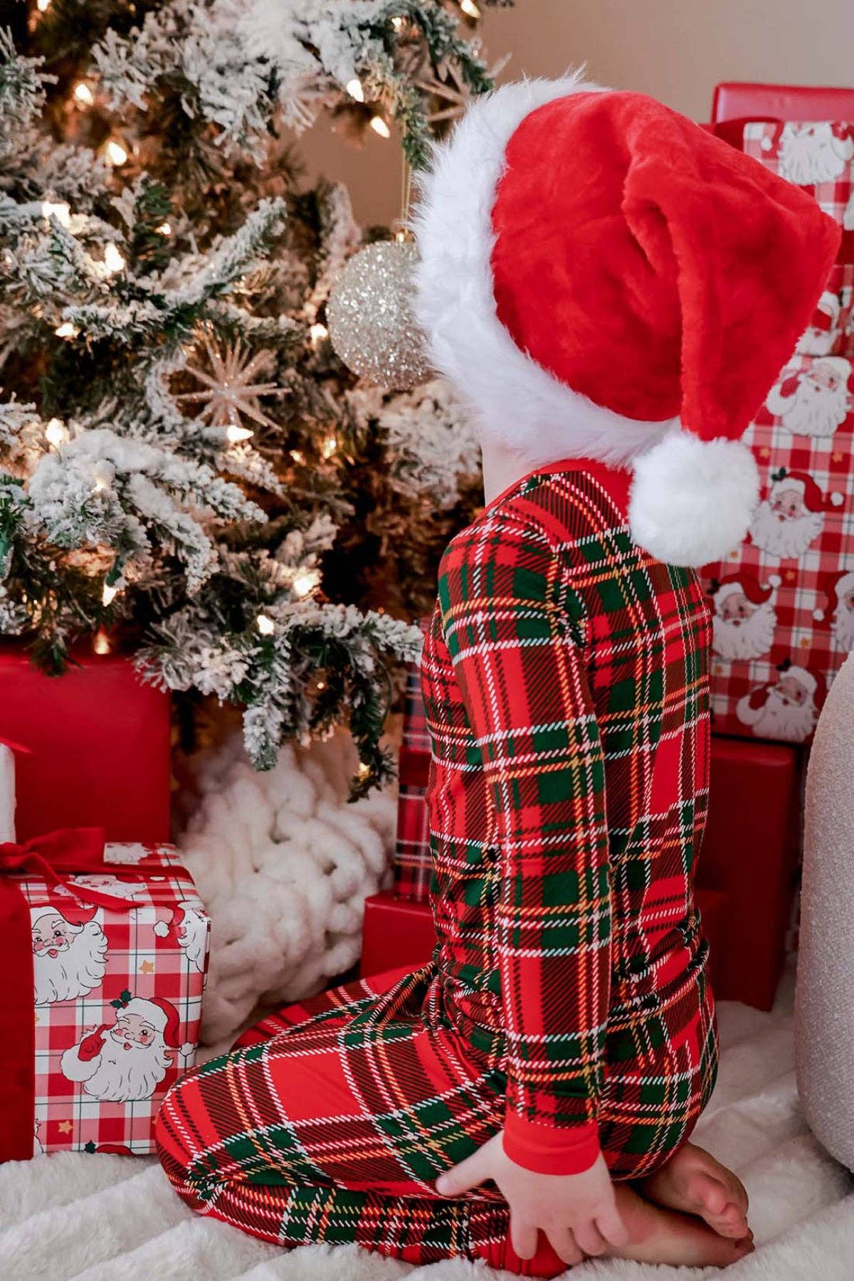 A child wearing red and green plaid pajamas and a Santa hat sits in front of a decorated Christmas tree with wrapped presents nearby.