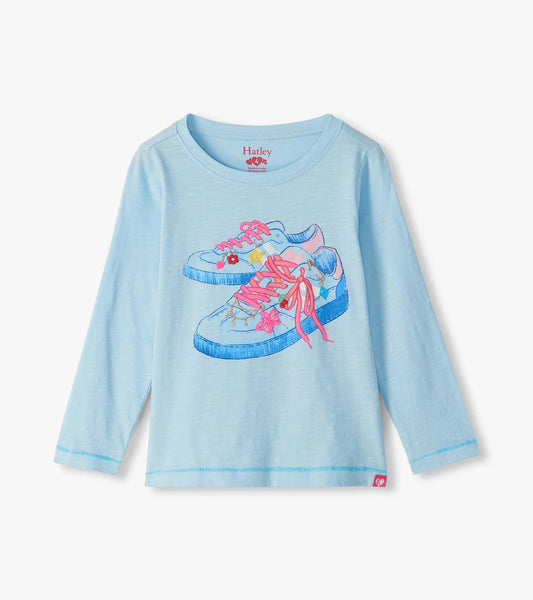 A light blue long-sleeve t-shirt for kids by Hurley, featuring a graphic print of two sneakers on the front with pink and blue details.